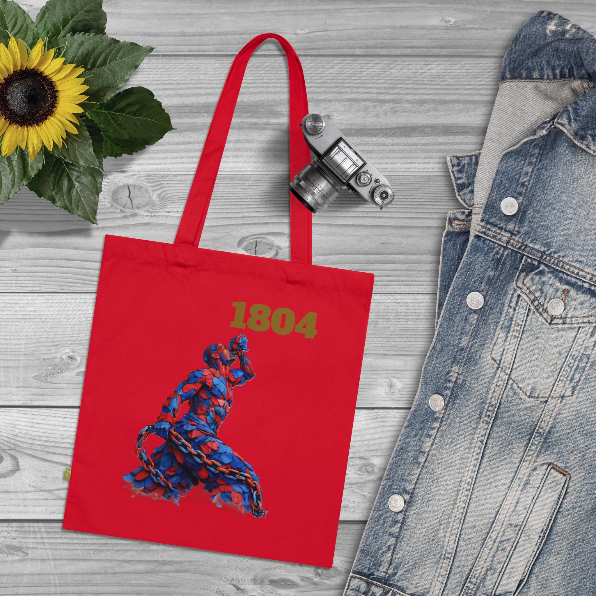 Red tote bag with '1804' and a dancer design, placed on a wooden surface with a sunflower and denim jacket.